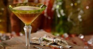 A martini glass filled with a layered cocktail sits on a rustic wooden surface. Several small paint tubes and brushes are scattered nearby. The background features colorful, blurred lights, creating a warm, artistic ambiance.