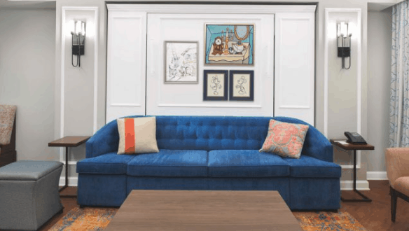 A Disney's Riviera Resort-themed living room with a blue sofa, Disney wall art, pillows, sconces, and a wood coffee table.