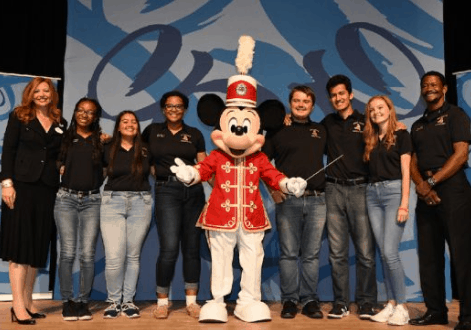 Mickey Mouse in a red band uniform joins a high school band and others on stage at Walt Disney World for a lively celebration.