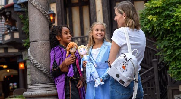 Young guests in Frozen princess dresses beam at a woman sporting an Olaf backpack, surrounded by Frozen 2 merchandise at Disney Parks.