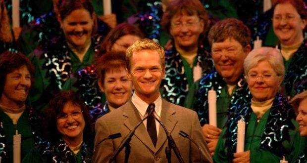 Neil Patrick Harris in a suit hosts Candlelight Processional, standing before a choir with candles and festive Epcot decorations behind them.