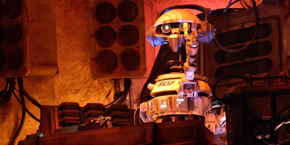 A robot with a dome-shaped head, glowing blue chest lights, and mechanical arms sits amidst speakers, wires, and equipment in a dimly lit, industrial sci-fi setting reminiscent of Star Wars: Galaxy's Edge Food markets.