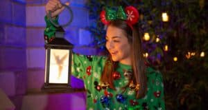 Disney fan wearing a festive sweater and Mickey ears beams, holding a Tinker Bell lantern at Mickey’s Very Merry Christmas Party.