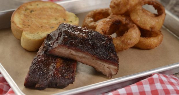Barbecue ribs, onion rings, and garlic bread served at Regal Eagle Smokehouse in Epcot on a red checkered tray.