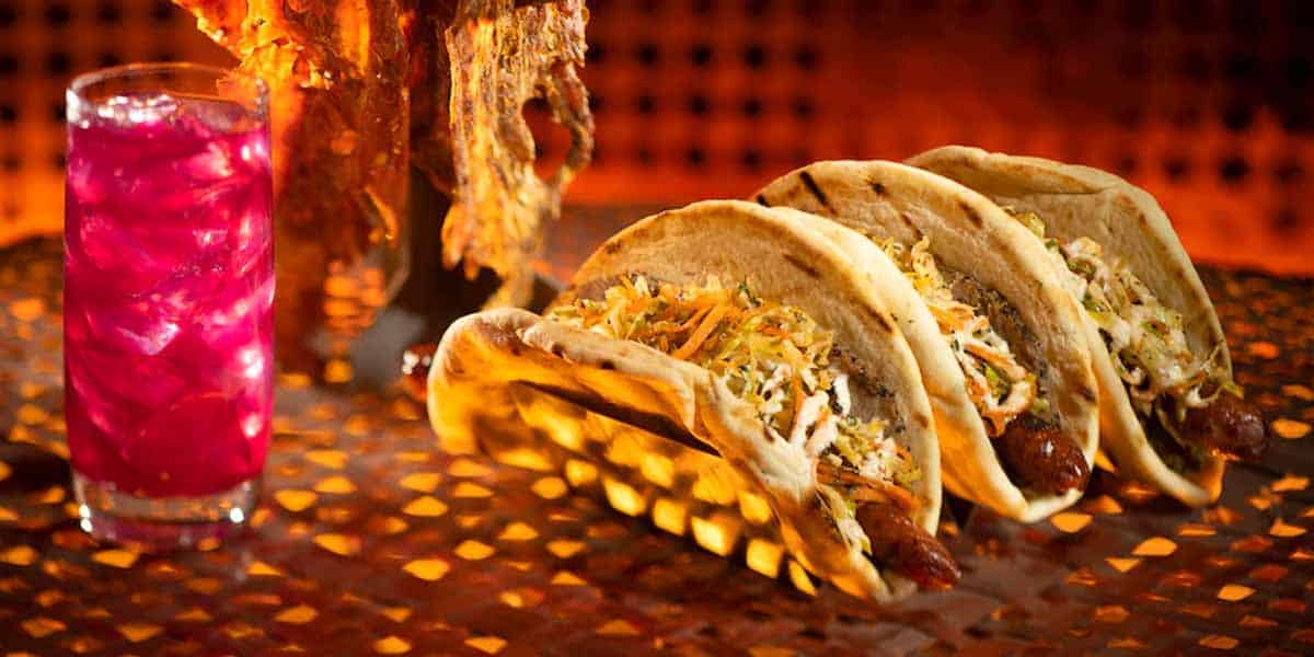 Three tacos filled with shredded vegetables and meat are lined up on a metal tray—just like a meal from Star Wars: Galaxy's Edge Food. A glass of pink iced drink sits to the left, with golden, warmly lit tones in the background.