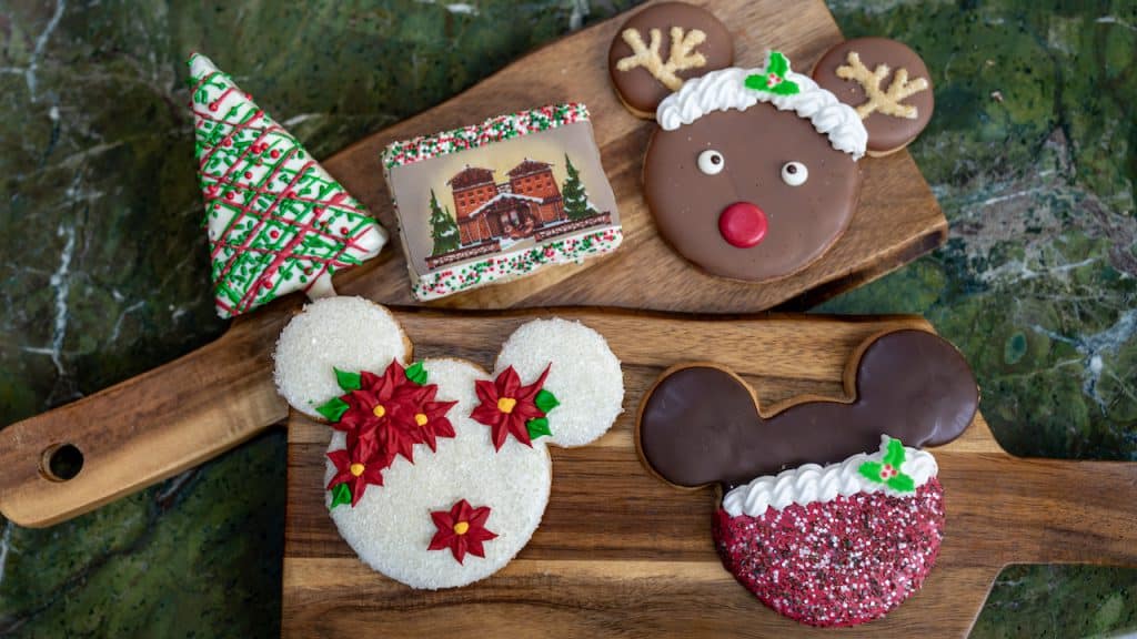 Five Disney-themed holiday cookies—Mickey shapes, tree, gingerbread house—on wooden boards, a treat for December park-goers.