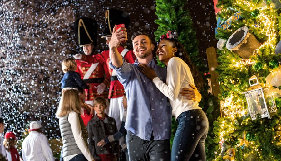 Guests snap a selfie in falling snow by the Disney Springs Christmas Tree, immersed in the trail’s festive holiday magic.