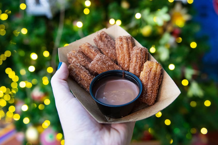 Churros with chocolate dip held up on the Disney Springs Christmas Tree Trail, surrounded by sparkling holiday lights at Disney World.