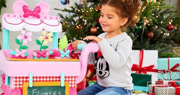 A happy child enjoys a shopDisney pink flower stand toy near a sparkling Christmas tree and festive Disney holiday presents.