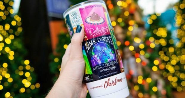 A festive holiday cup is held up with sparkling Christmas lights and elaborately decorated trees glowing in a theme park setting.