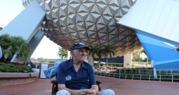 A WWII veteran in a wheelchair enjoys the view of Epcot’s Spaceship Earth, surrounded by Disney World’s lively atmosphere.