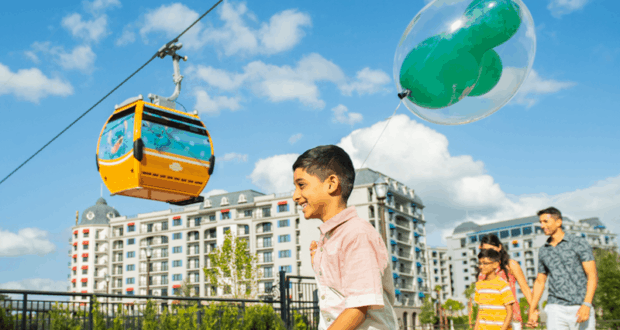 Young guest with a balloon strolls by Disney Skyliner and resort towers, enjoying the sunny excitement of Walt Disney World.