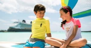 Two happy kids relax on a sandy beach as the Disney Cruise Line ship and a sailboat glide by under a bright, sunny sky.
