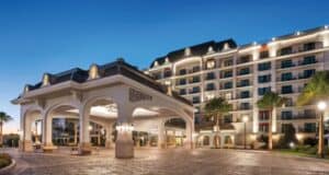The image shows a luxurious hotel entrance with an ornate, covered driveway and a grand multi-story building in the background. The scene is lit warmly against the evening sky, flanked by palm trees and soft, ambient lights.
