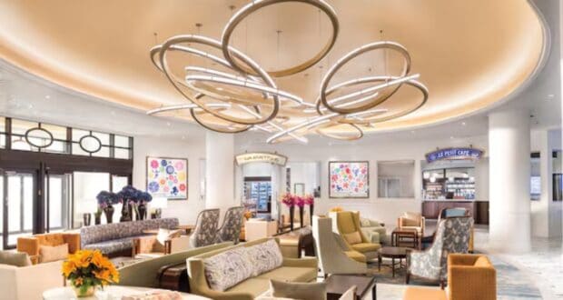 Elegant hotel lobby with modern circular chandeliers, comfortable seating, and floral artwork. The space is bright and inviting, featuring a small café area with beverages.