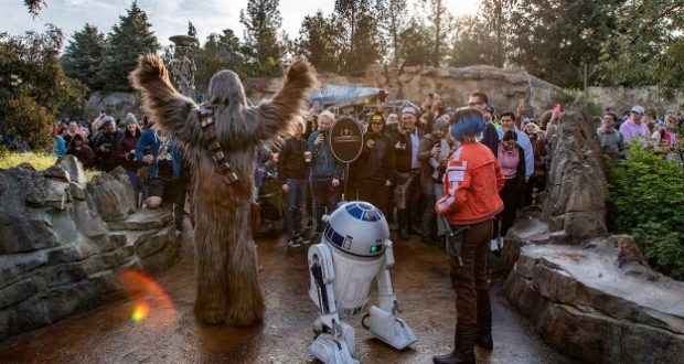 Chewbacca and R2-D2 welcome fans in Star Wars: Galaxy’s Edge at Disneyland, with Batuu’s rocky landscape and trees behind them.