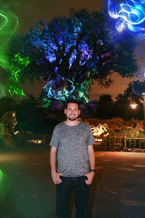 A smiling guest enjoys a dazzling, neon-lit Tree of Life at Walt Disney World’s 15th Anniversary PhotoPass celebration.
