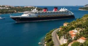 Disney Cruise Line ship glides by a scenic Greek town, red rooftops and green hills evoking the spirit of Disney adventures.