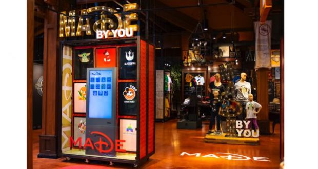 A MADE kiosk in a Disney park store, showcasing personalized Disney shirts, with mannequins and apparel in the background.