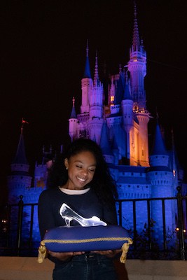 A guest presents a sparkling glass slipper on a cushion in front of Cinderella Castle, glowing at night—a magical PhotoPass!.
