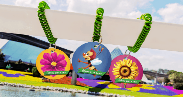 Disney Gift Cards with playful flower and bee art dangle from green spirals amid Epcot gardens, celebrating FreshEpcot vibes.