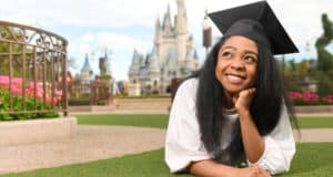 Graduate in cap relaxes on the grass near Cinderella Castle, celebrating Magic Kingdom graduation under sunny skies.