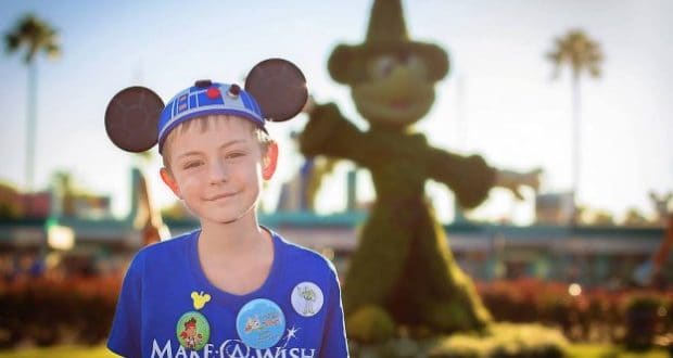 Excited young guest at Disney parks in a Star Wars Mickey hat celebrates World Wish Day near Sorcerer Mickey topiary.