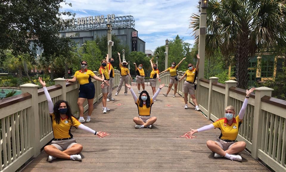 Disney Springs Cast Members in yellow shirts celebrate together on the bridge, masked and socially distanced during reopening.