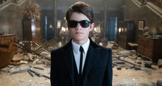 Artemis Fowl, clad in a sleek black suit and shades, stands among wreckage—inviting fans into his thrilling Disney+ world.