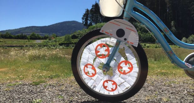 Blue bike’s front wheel decorated with Star Wars paper crafts, featuring BB-8, shines under sunny skies with mountain views.