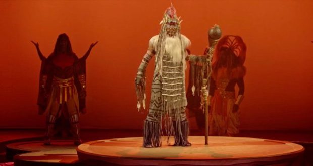 A Lion King performer in stunning tribal costume wows the stage at Disneyland Paris, staff in hand against a vivid orange backdrop.