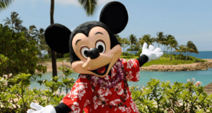 Mickey Mouse greets fans in a vibrant red Hawaiian shirt by palm trees and the lagoon at Disney’s Aulani Resort in Hawaii.