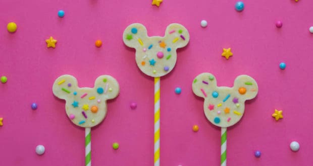 Mickey-shaped lollipops with colorful sprinkles pop against a pink, starry Disney-inspired background, perfect for park festivities.