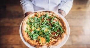 A person holding a freshly baked pizza topped with arugula and assorted vegetables on a thin, crispy crust, standing on a wooden floor. The pizza is presented on a white plate.