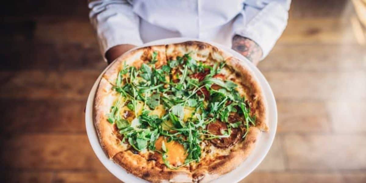 A person holding a freshly baked pizza topped with arugula and assorted vegetables on a thin, crispy crust, standing on a wooden floor. The pizza is presented on a white plate.
