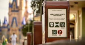 Trash can with Disney World health and safety sign, guest’s park bag visible, Cinderella Castle softly blurred in the distance.