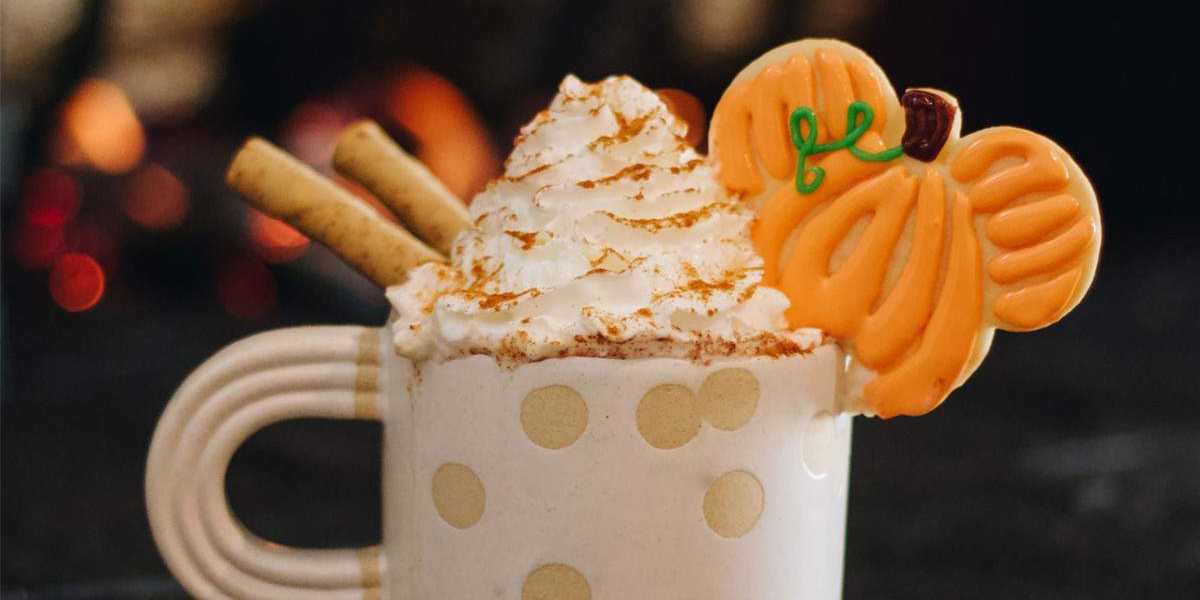 A cozy white mug with polka dots is filled with a swirl of whipped cream, topped with a dusting of cinnamon, two rolled wafer cookies, and a pumpkin-shaped cookie. The background is softly blurred with warm lights.