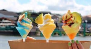Three colorful ice cream cones with unique toppings are held up against a waterfront backdrop. The ice creams feature blue, yellow, and orange swirls topped with cookies, fruit slices, and drizzle, respectively. The background shows a calm water body and buildings.