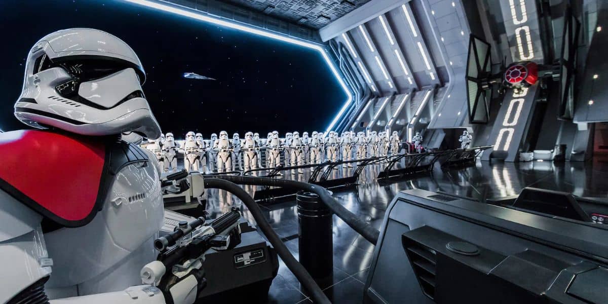 A group of stormtroopers stands in formation inside a futuristic spacecraft. One stormtrooper with a red shoulder pad is in the foreground. A large window shows a view of space with stars and a distant spacecraft.