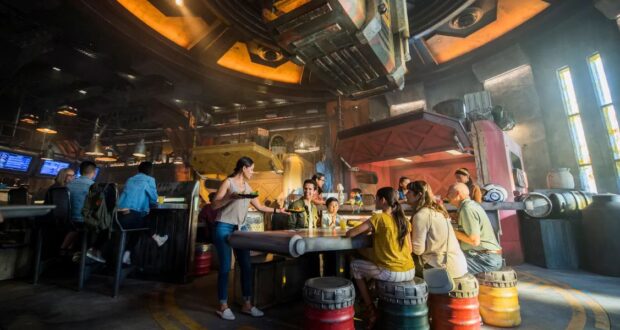 A group of people sit around a futuristic, circular table in a lively, sci-fi themed bar. The decor includes metallic elements, colorful lights, and industrial designs. A server carries a tray with drinks, and scattered barrels add to the ambiance.
