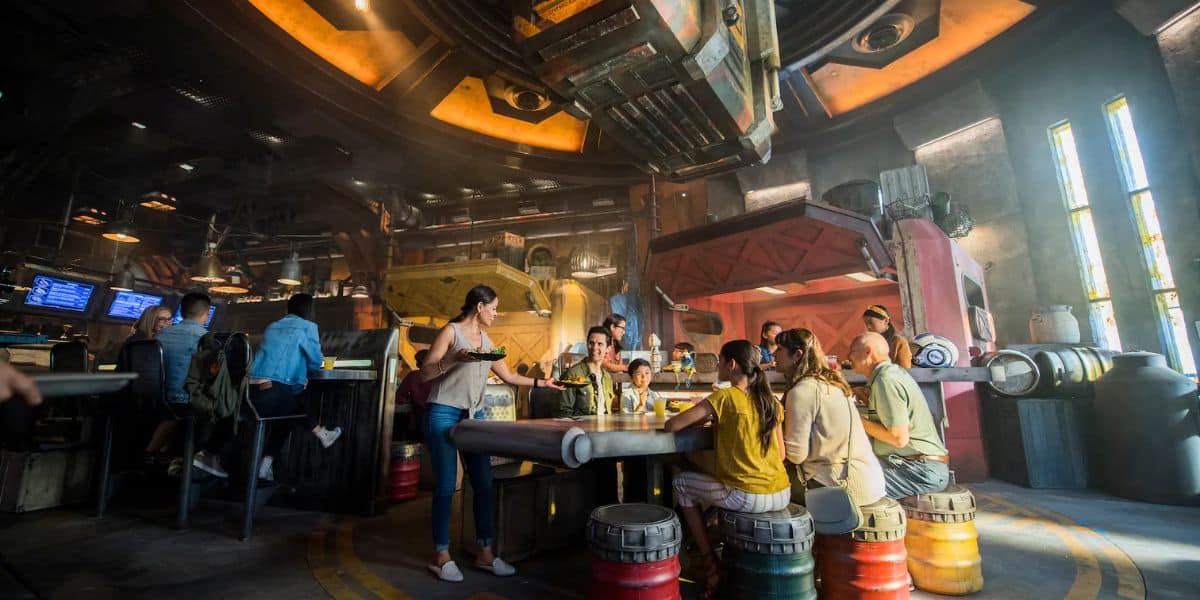 A group of people sit around a futuristic, circular table in a lively, sci-fi themed bar. The decor includes metallic elements, colorful lights, and industrial designs. A server carries a tray with drinks, and scattered barrels add to the ambiance.