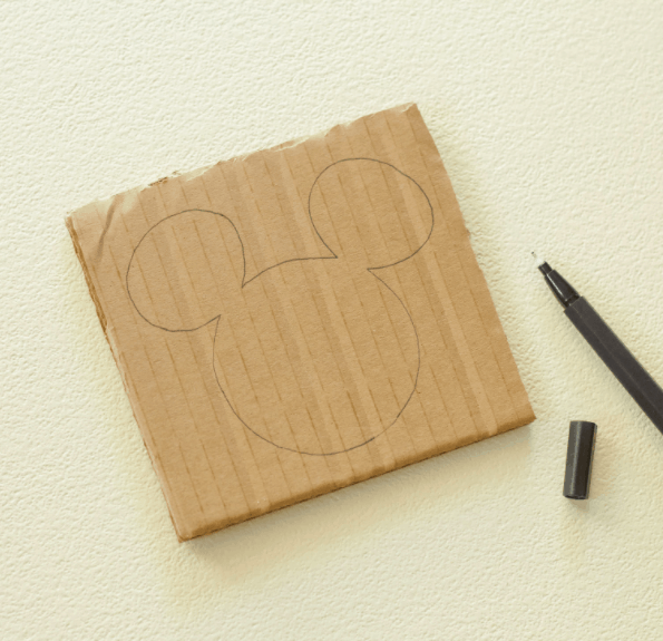 A square piece of cardboard featuring an iconic Mickey Mouse head outline, perfect for Disney-inspired DIY projects, rests beside an uncapped black marker—ready for fans to create their own magical Mickey Yarn Toppers at home.