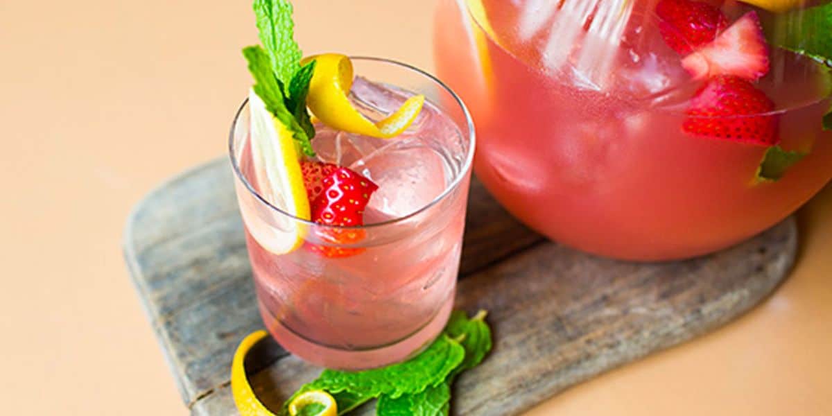 A refreshing pink drink in a glass garnished with mint leaves, a lemon twist, and sliced strawberries. The glass sits on a wooden board, with a larger pitcher of the same drink in the background. Lemon peel and mint are scattered around.