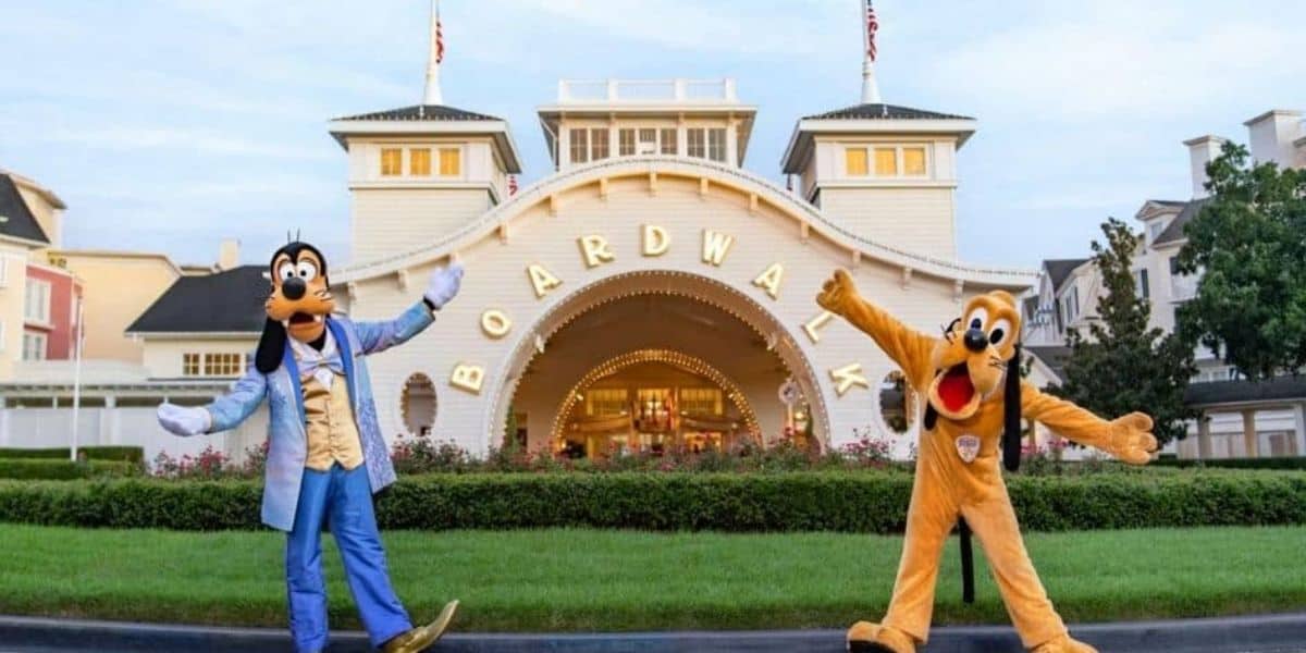Goofy and Pluto are posing cheerfully in front of the BoardWalk Inn entrance. The backdrop features the archway with "Boardwalk" prominently displayed, surrounded by charming, classic resort architecture and manicured greenery.