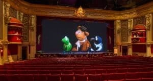 An ornate theater with red seats faces a large screen showing three puppet characters. The intricately designed interior features golden accents and red curtains. The theater is empty, and the screen displays a scene from a puppet show.