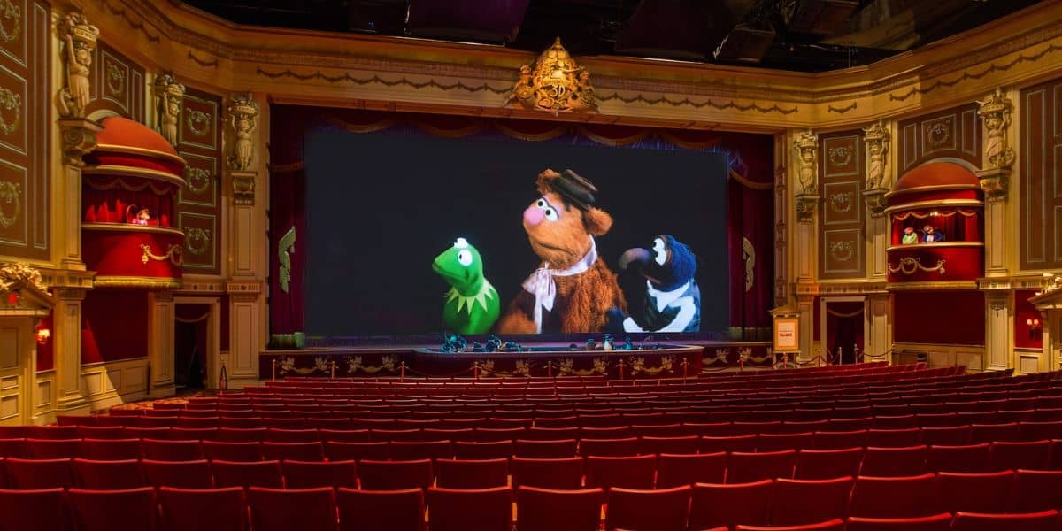 An ornate theater with red seats faces a large screen showing three puppet characters. The intricately designed interior features golden accents and red curtains. The theater is empty, and the screen displays a scene from a puppet show.