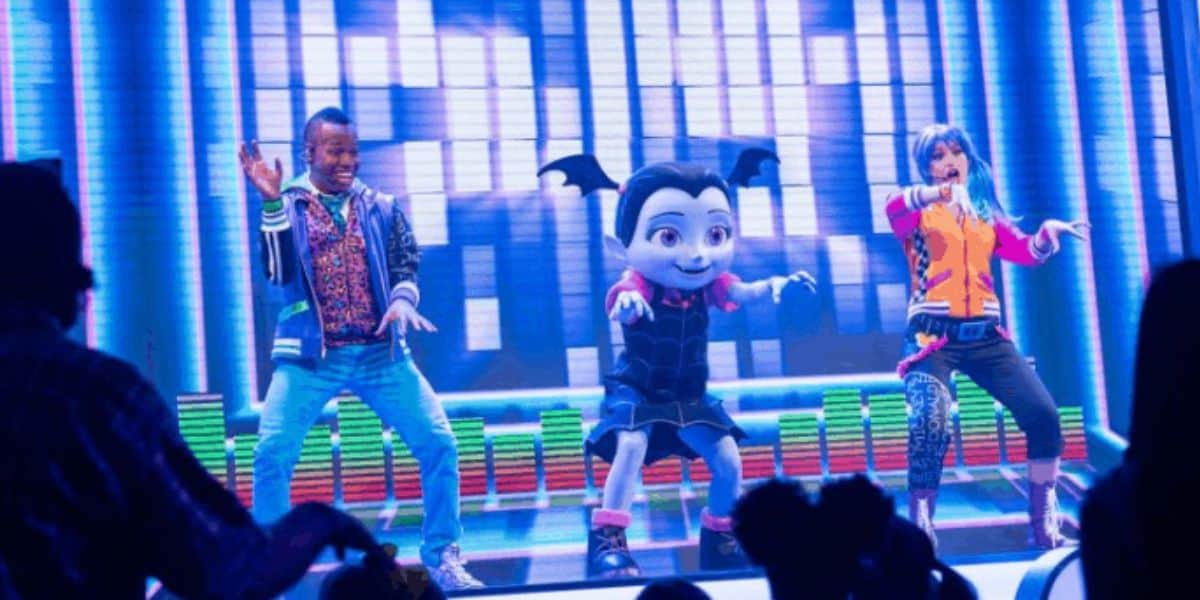 A person in colorful clothing, a costumed character resembling a vampire with bat-like ears, and another person with blue hair and an orange jacket dance energetically on a stage with a vibrant, digital backdrop.