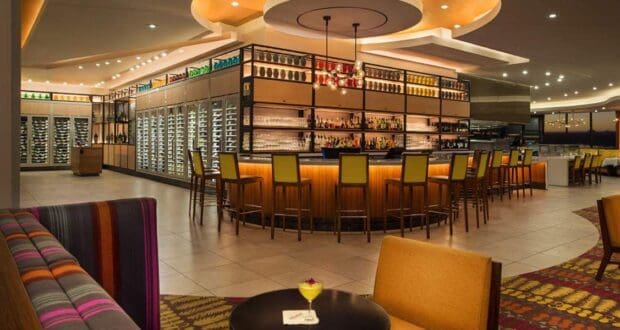 A modern bar with a circular counter surrounded by yellow stools. The walls are lined with shelves filled with bottles. The space is lit by warm, ceiling-mounted lights. A small table with a drink and flower is in the foreground.