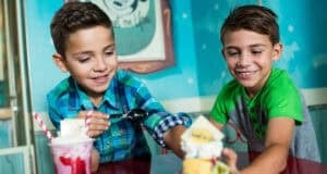 Two young boys sit at a table, smiling and enjoying ice cream sundaes while chatting about their unpopular Disney World opinions. One boy scoops ice cream from a glass with whipped cream and straws; the other holds a sundae with a cookie and yellow topping.