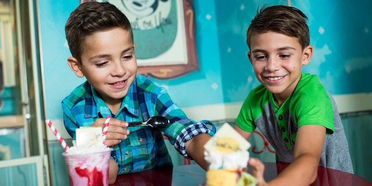 Two young boys sit at a table, smiling and enjoying ice cream sundaes while chatting about their unpopular Disney World opinions. One boy scoops ice cream from a glass with whipped cream and straws; the other holds a sundae with a cookie and yellow topping.